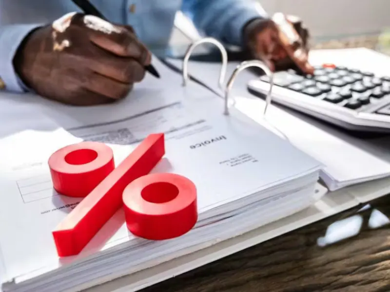 Person writing notes with a pen on an invoice, calculator nearby, and large red percentage symbol, representing financial calculations and business services.